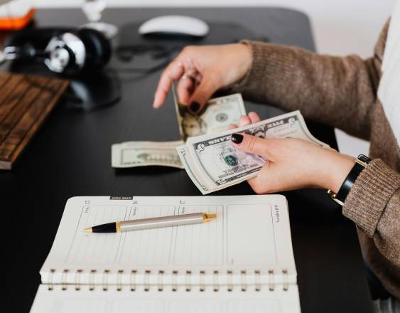 PEXELS | Una mujer contando dinero sobre un escritorio. Realiza apuntes para llevar un registro.